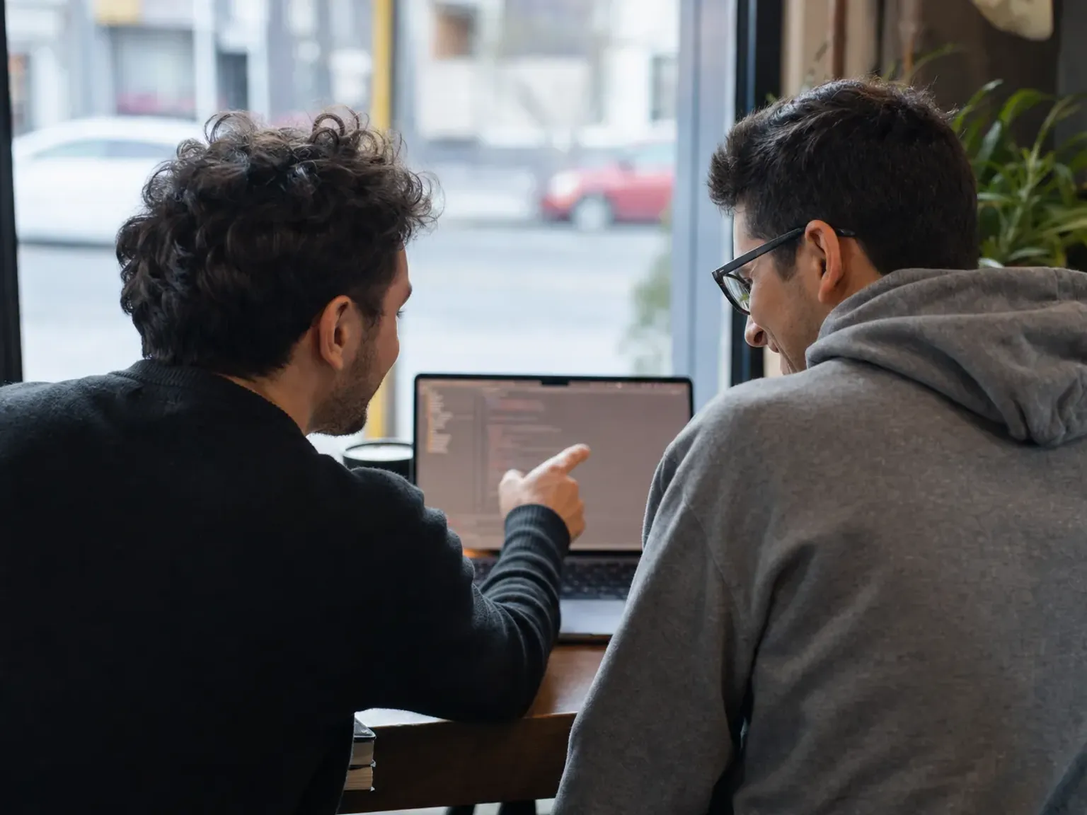Two teammates pairing at a single laptop in a relaxed workspace corner, one pointing at the screen mid-explanation.