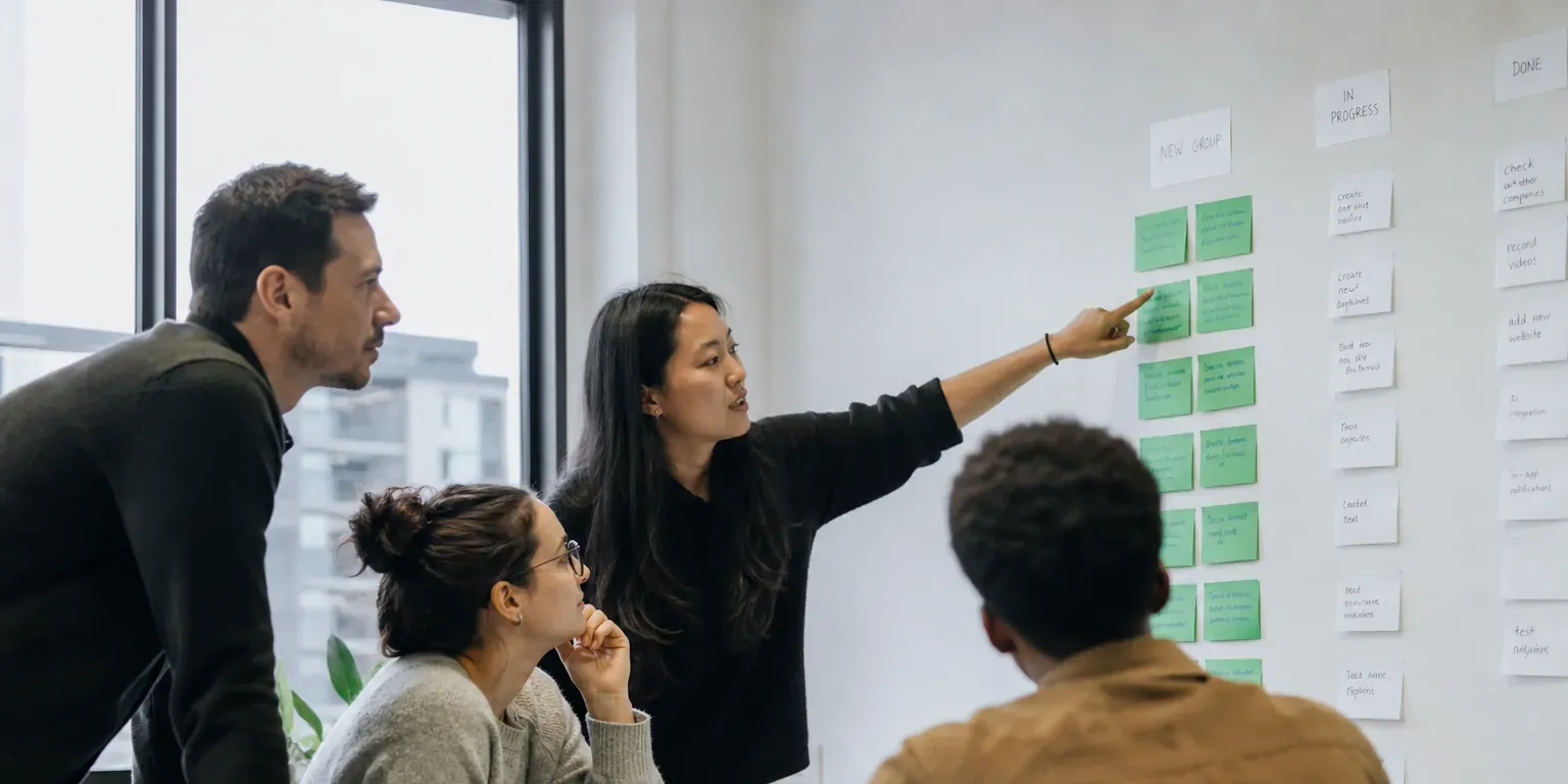 A cross-functional product team gathered around a laptop and a wall of sticky notes, reviewing research findings together.